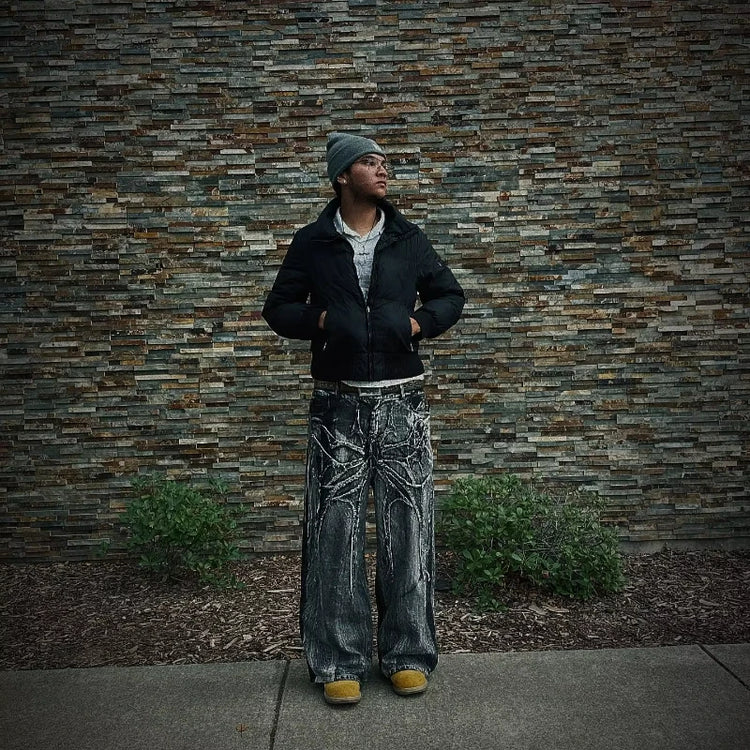 Man wearing retro spider phantom pants with black jacket and gray beanie standing against stone wall